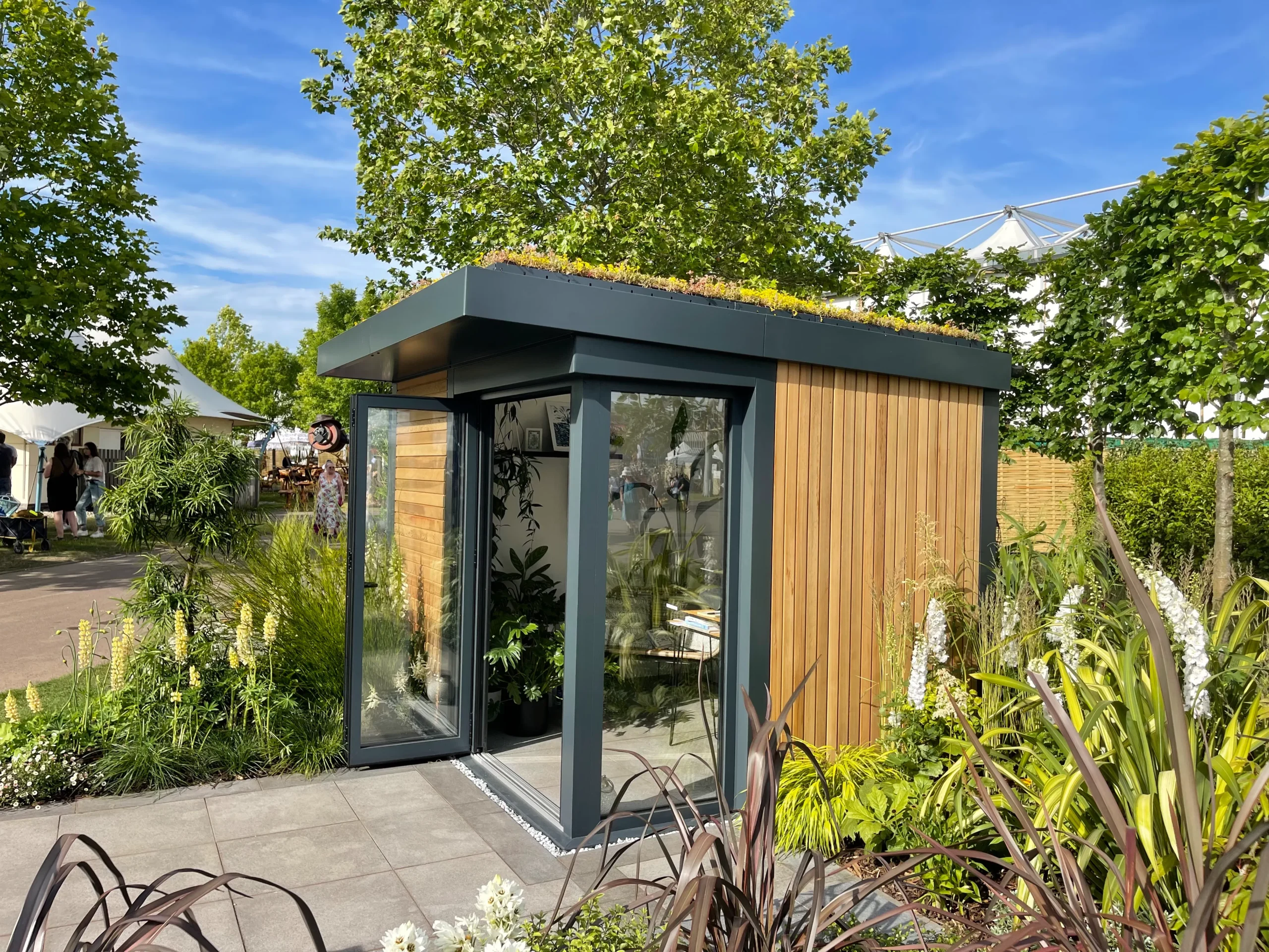 A modern garden building with a flat, green roof, wood-clad walls, and glass doors, set amidst lush landscaping of various plants and flowers on a paved patio, with a bright blue sky and trees in the background.