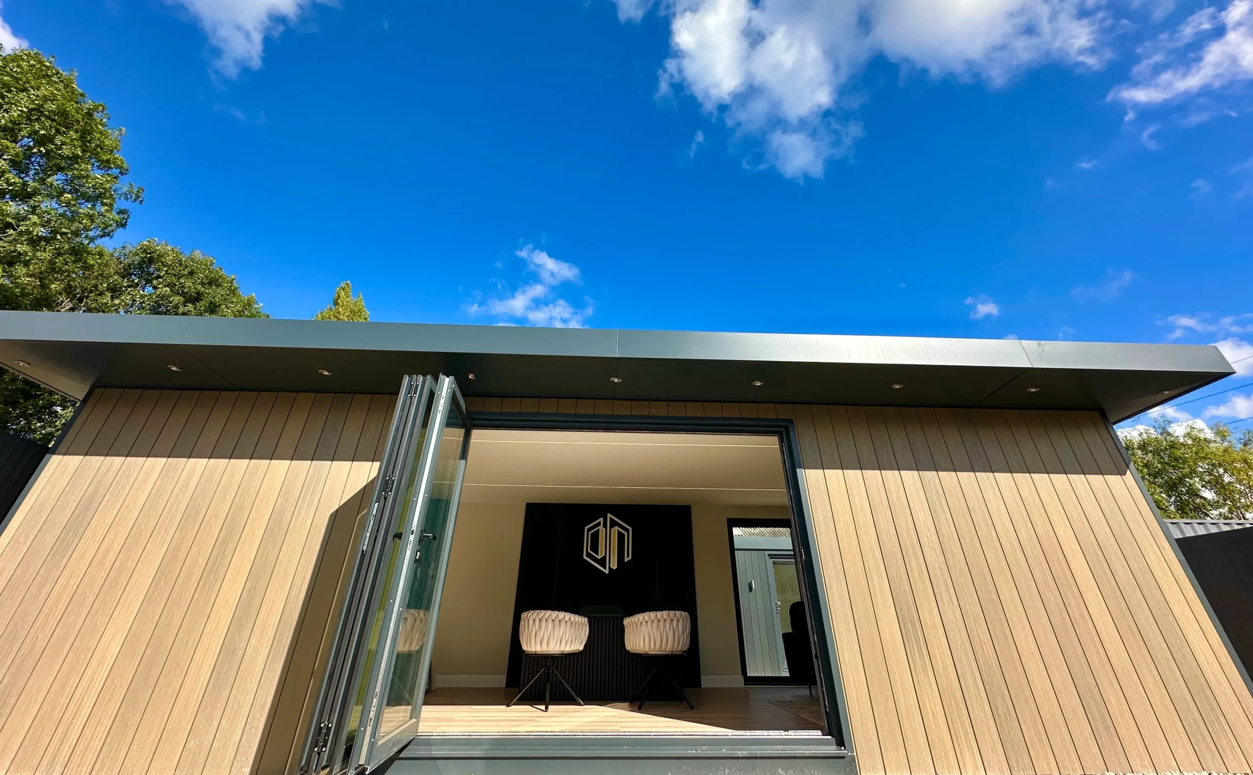 Exterior view of a modern building with a wooden facade. Large, open glass doors reveal an interior with two chairs and a logo on the back wall. The sky is bright blue with scattered clouds. The building has a dark, flat roof with recessed lighting. Trees are visible around the structure.