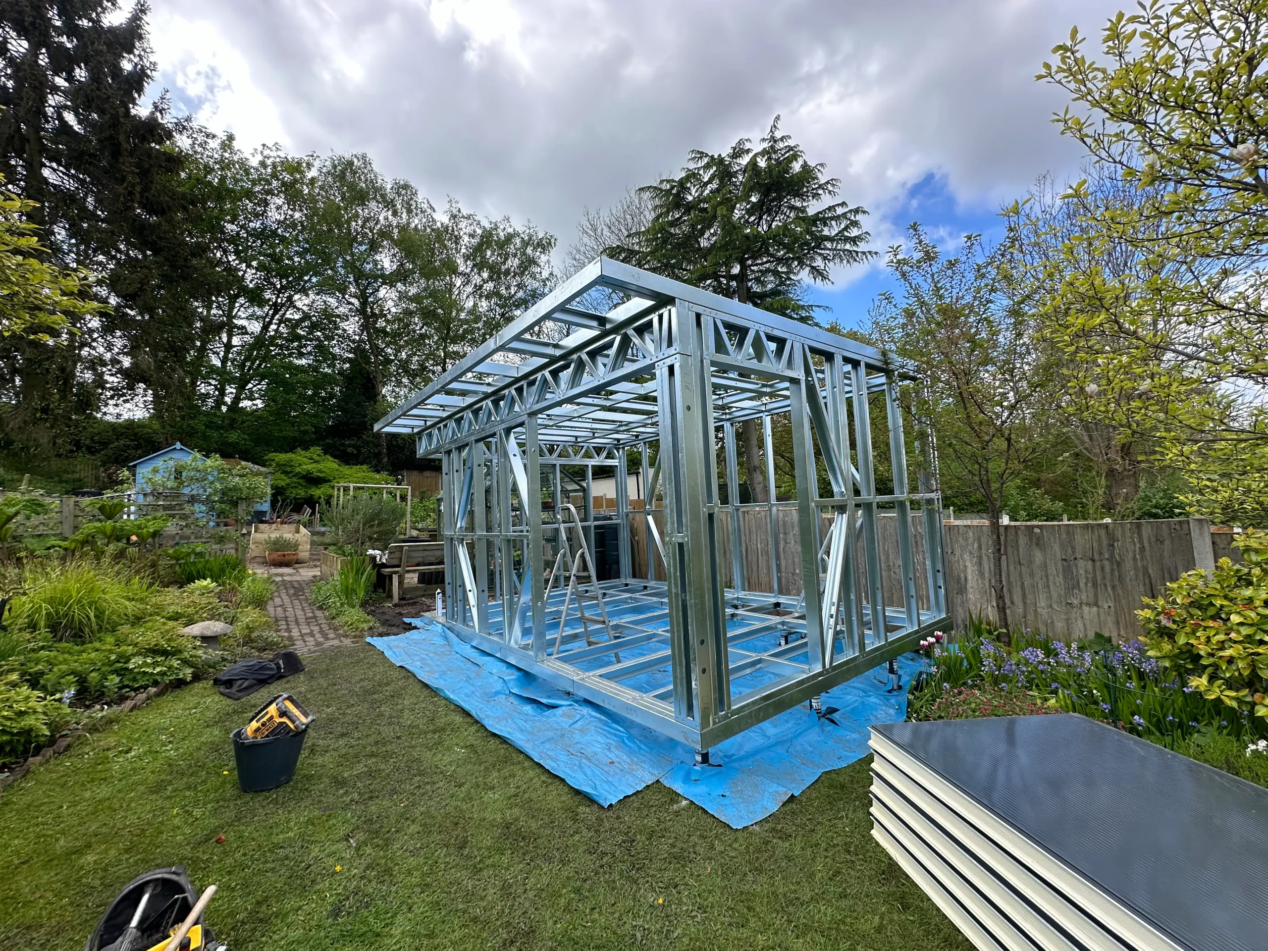 Metal frame of a small building under construction, set on a blue tarp on a lawn. A stack of insulated panels rests to the side. The sky is cloudy with hints of blue, and trees and shrubbery surround the construction site. Construction tools are nearby.