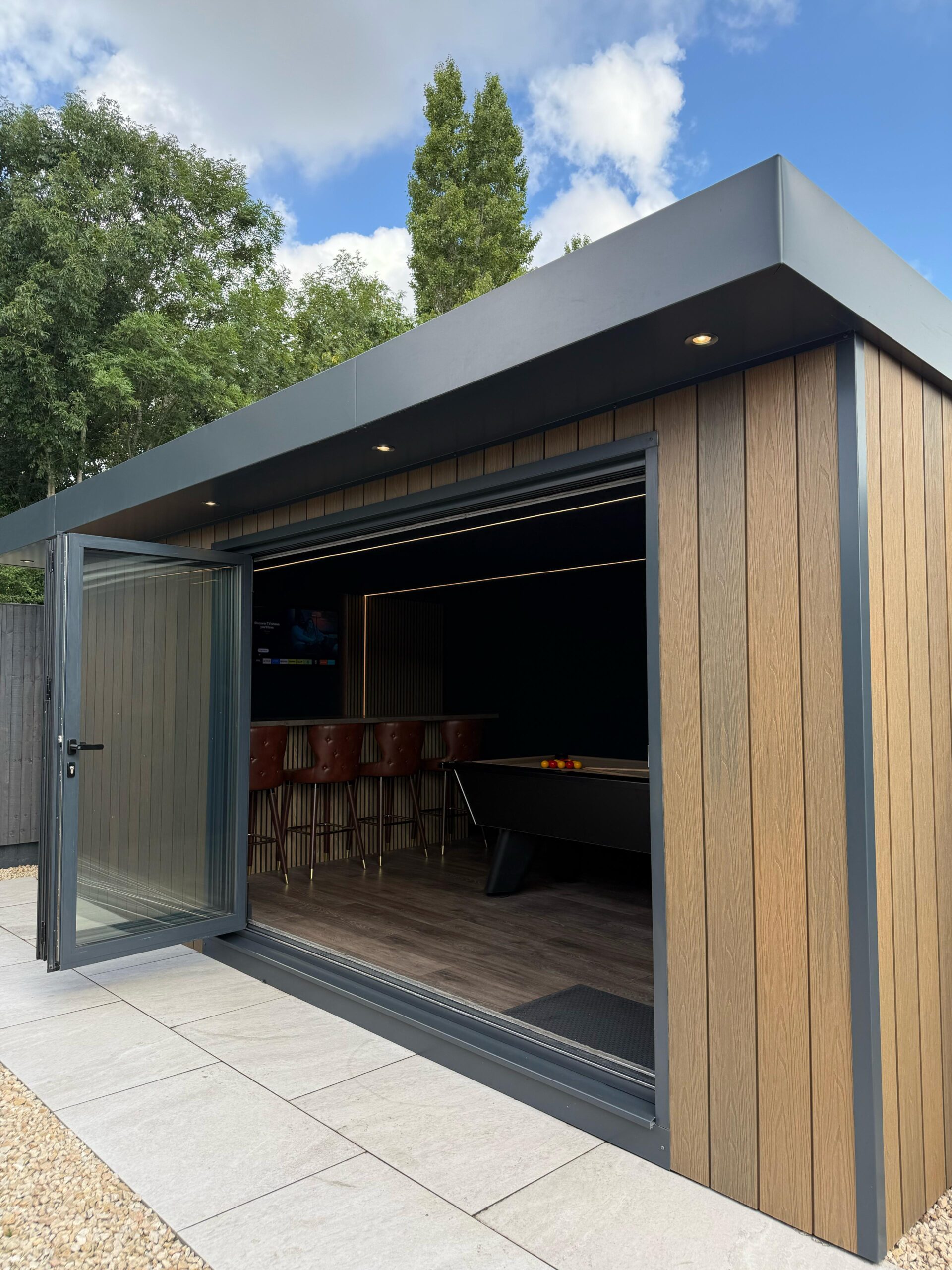 A modern outdoor entertainment space with a pool table, bar, and seating, featuring bi-fold doors, wooden paneling, and a flat roof under a partly cloudy blue sky.
